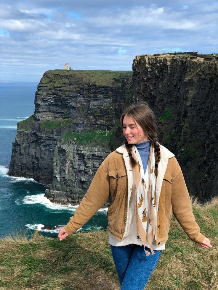 a woman standing on top of a grass covered hillside next to the ocean and cliffs