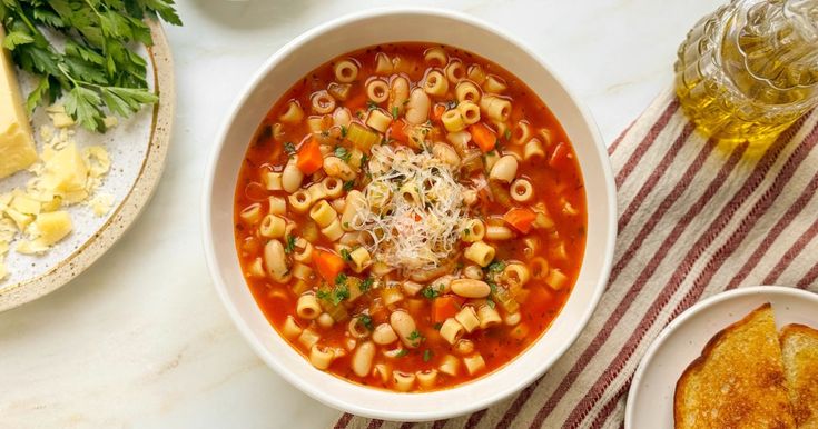 a bowl of pasta soup on a table with bread and parmesan cheeses