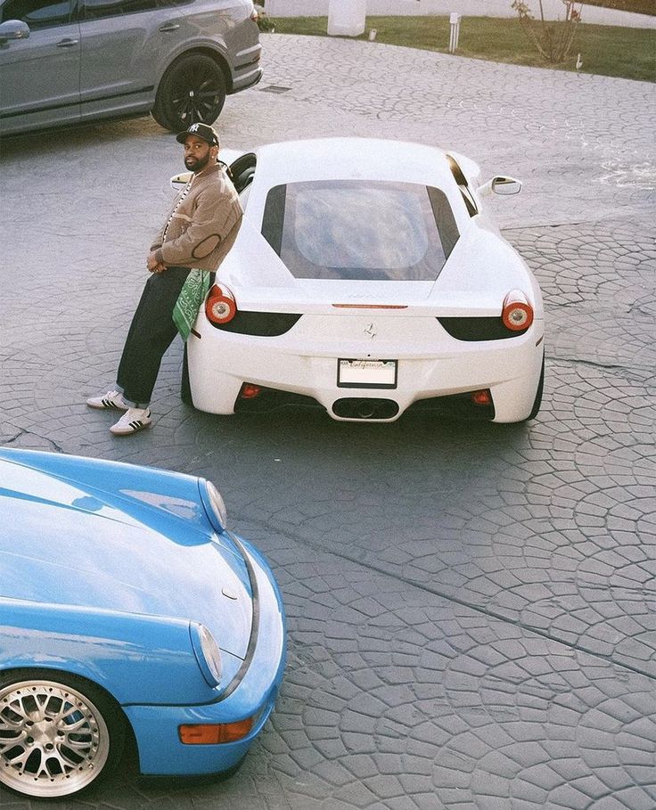 a man sitting on top of a white sports car next to another blue sports car