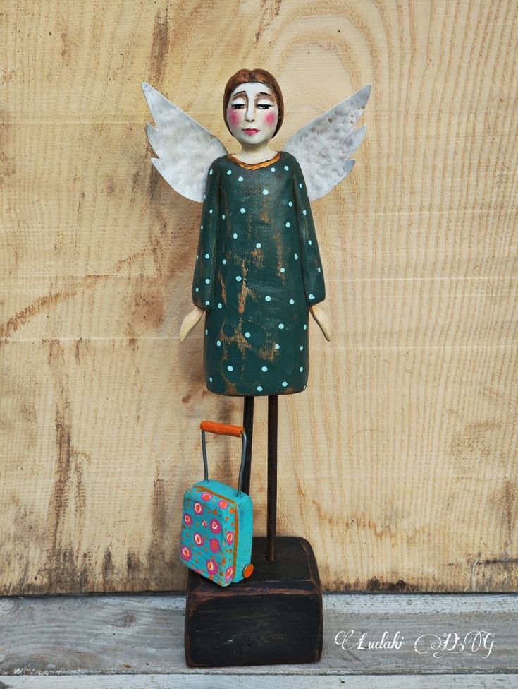 a wooden angel with green wings and polka dots holding a blue suitcase in front of a wood plank wall