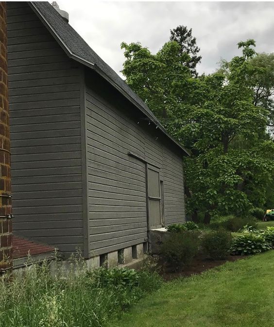 the side of a gray building with green grass and bushes around it, next to a brick wall