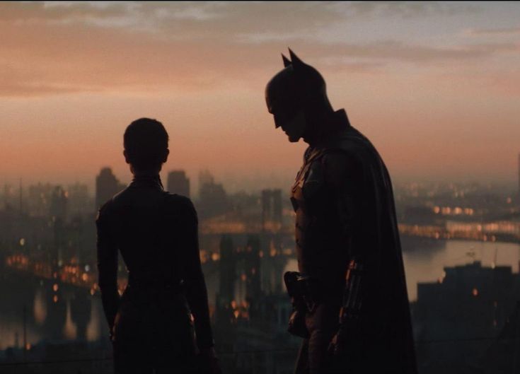 two people standing on top of a building looking at the city in the distance with batman and catwoman silhouetted against an orange sky