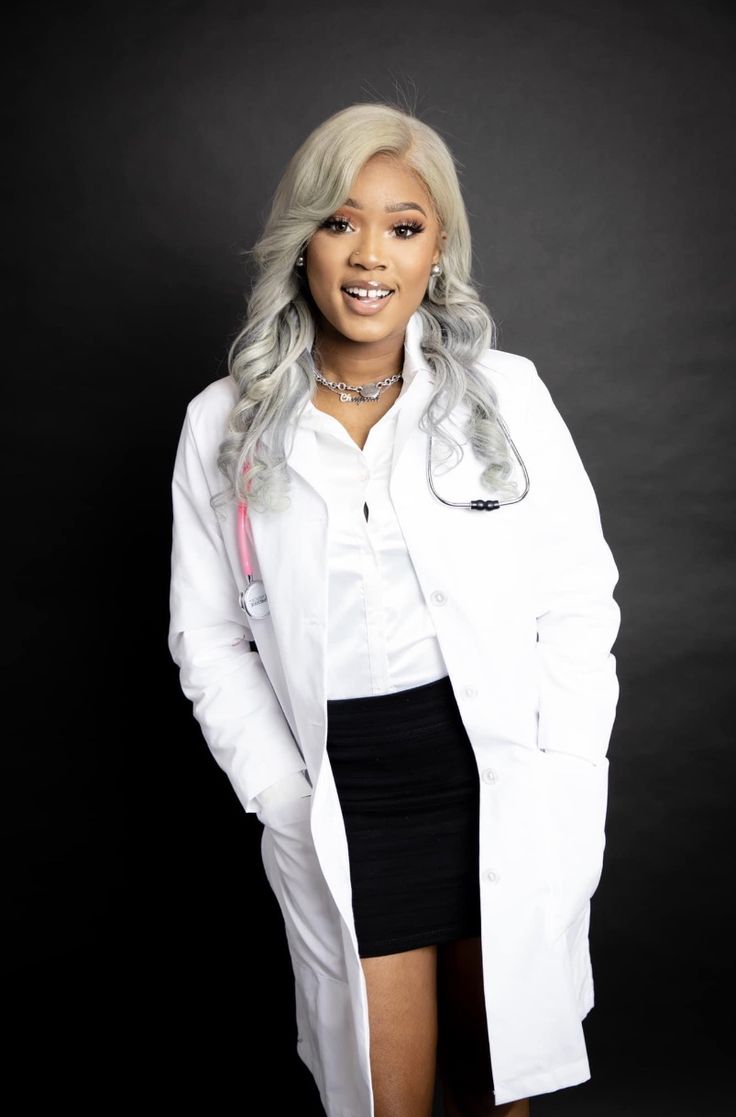 a woman with grey hair wearing a white lab coat and black skirt, standing in front of a dark background