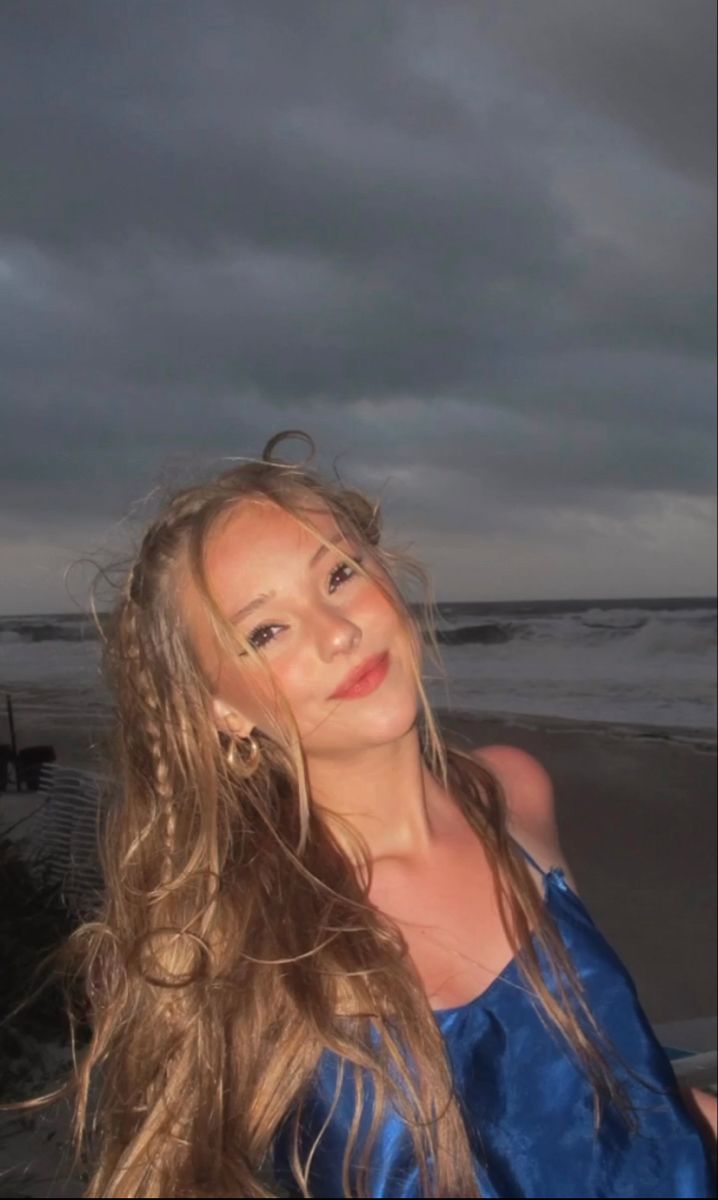 a beautiful young woman standing on top of a beach next to the ocean under a cloudy sky