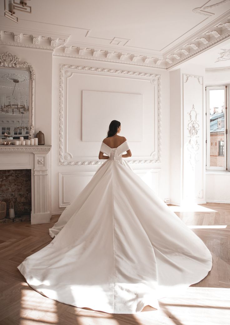 a woman in a white wedding dress is standing near a fireplace with her back to the camera