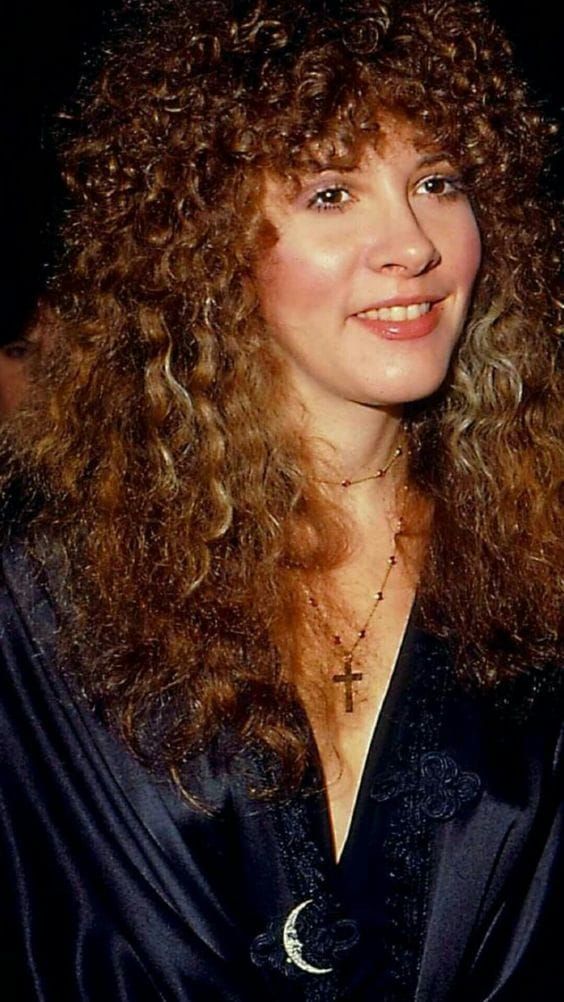 a close up of a person wearing a black shirt and long curly hair with a cross necklace