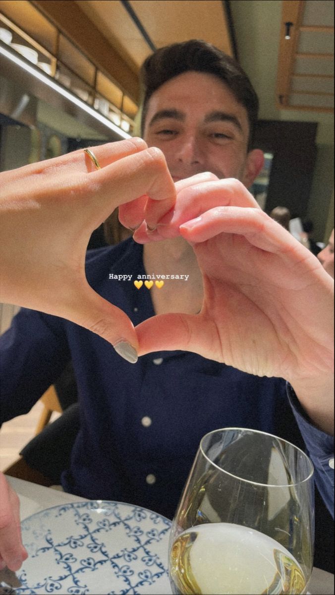 a man making a heart shape with his hands while sitting at a table in front of a wine glass