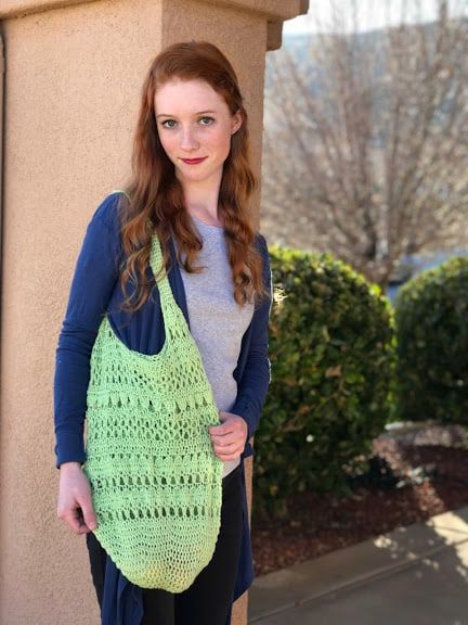 a woman holding a green crocheted bag on top of her shoulder and standing next to a building