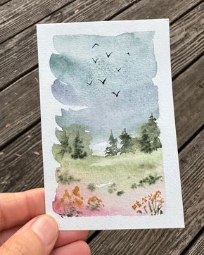 a person holding up a card with watercolors on it and birds flying above