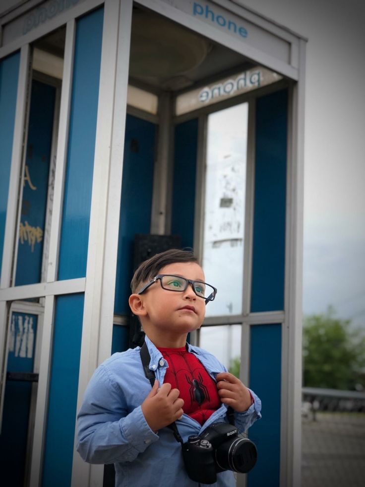 a young boy wearing glasses and holding a camera in front of a phone booth