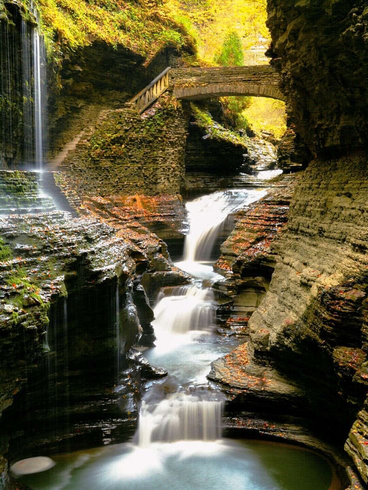 a waterfall with a bridge over it and water running down the side, in front of some rocks