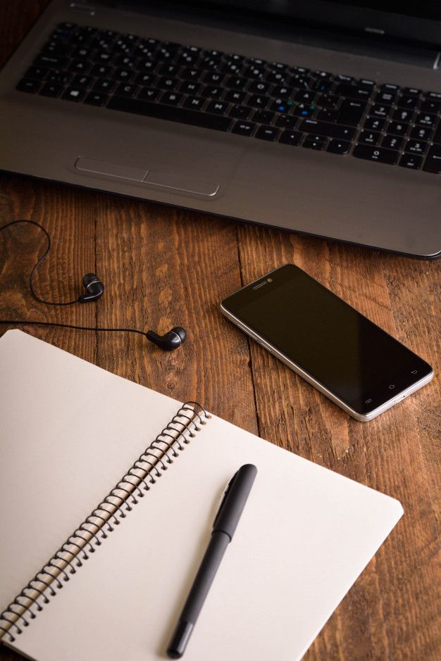 an open notebook, headphones and cell phone on a wooden table next to a laptop