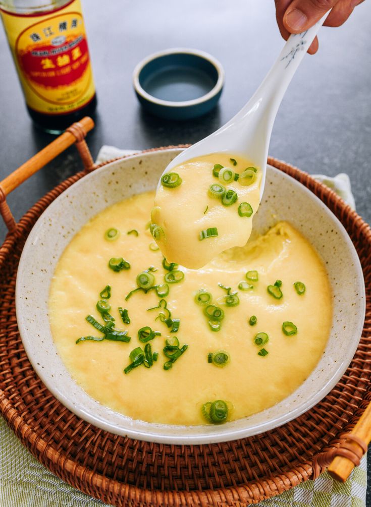 a person holding a spoon in a bowl filled with cheese and green onions on a table