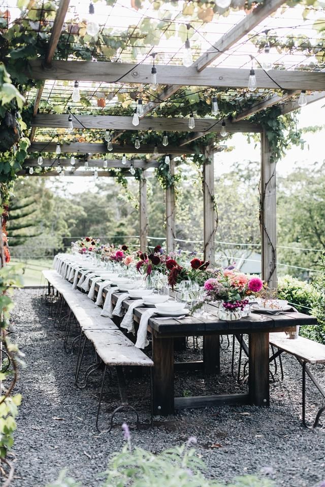 a long table with flowers on it under a pergolated roof in the middle of a garden