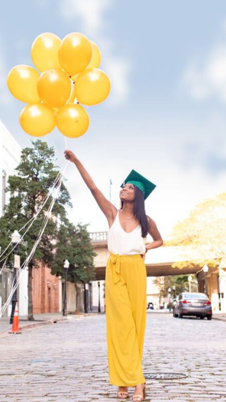 a woman in yellow pants and graduation cap holds up balloons while standing on the street