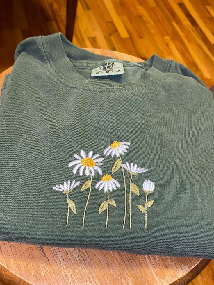 a green t - shirt with daisies embroidered on the front sitting on a wooden chair