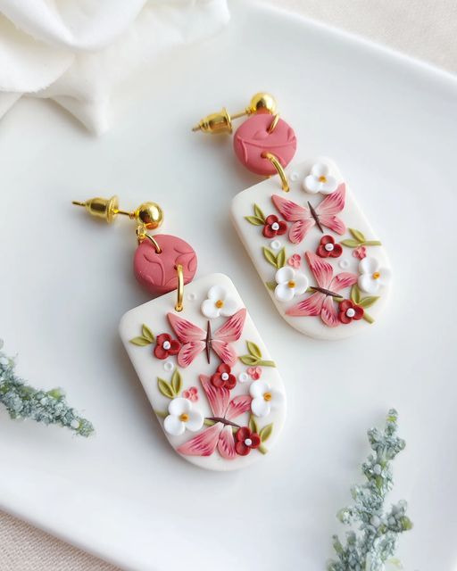 two white and pink flowered earrings on a plate next to some green plants with flowers