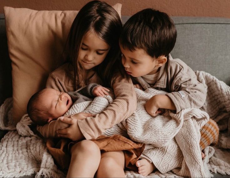 two young children sitting on a couch with their newborn babies in the arms and looking at each other