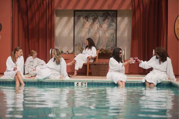 four women in white robes sitting on the edge of a swimming pool and drinking wine