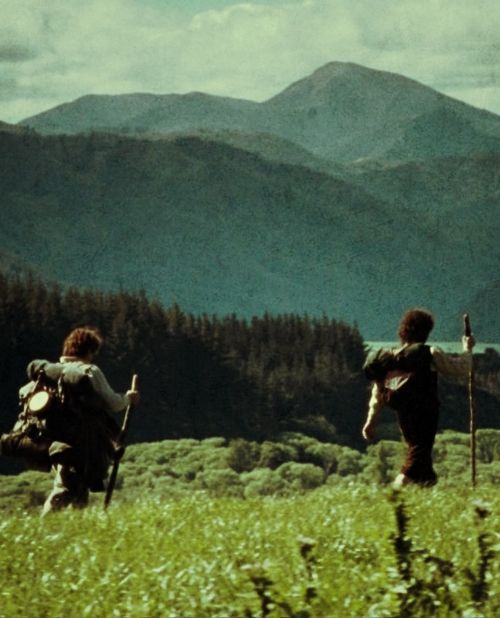 two people with backpacks are walking through the grass in front of some mountains and trees