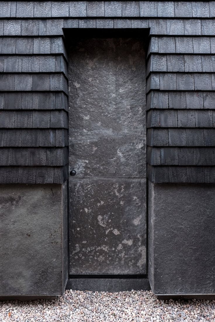 an open door on the side of a black shingled building with gravel around it