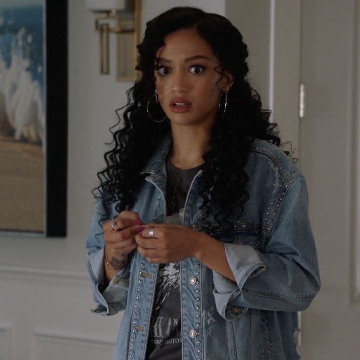 a woman with curly hair wearing a jean jacket and looking at the camera while holding a cell phone