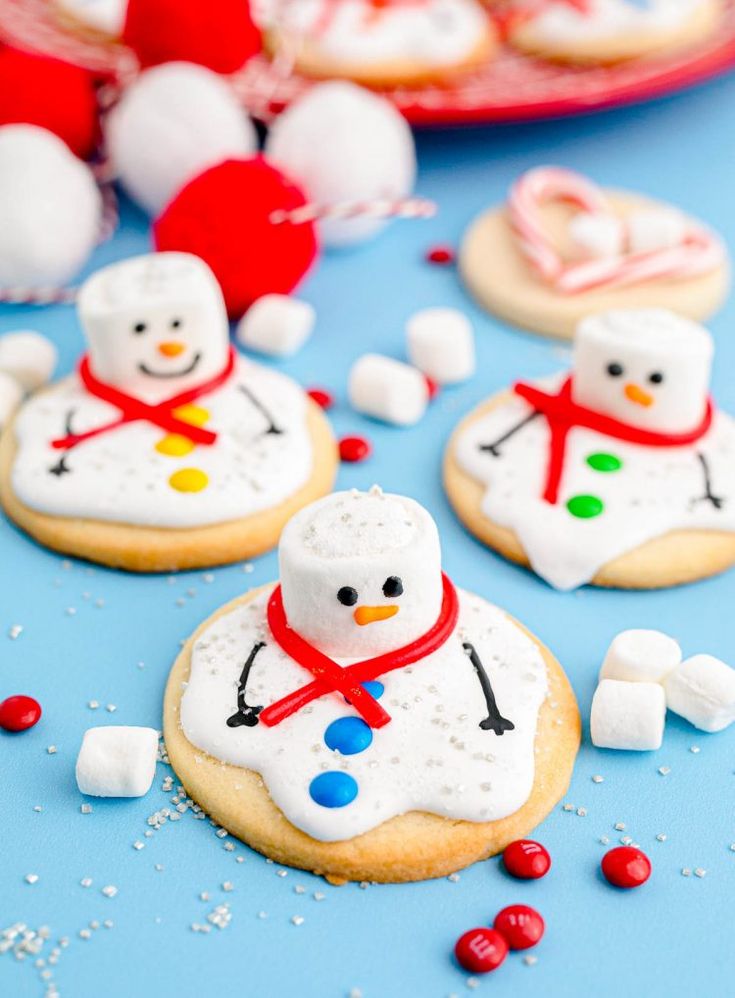 decorated cookies with marshmallows and snowmen on a blue tablecloth surrounded by other holiday treats