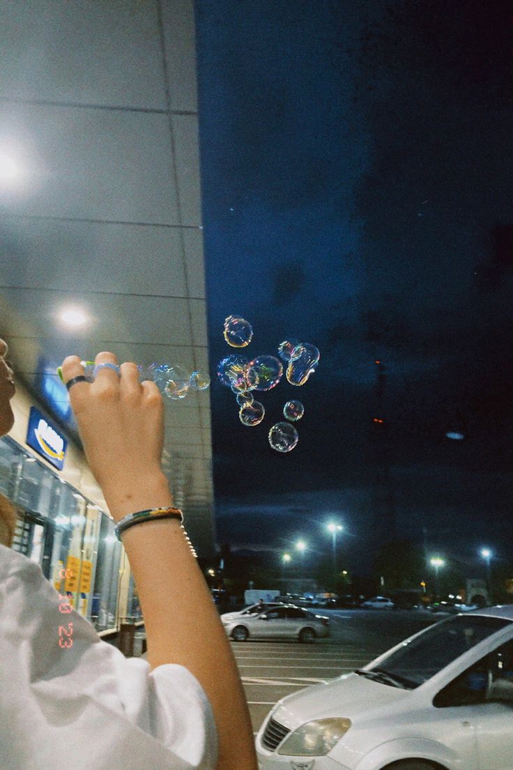 a woman blowing bubbles in front of a white car on a city street at night