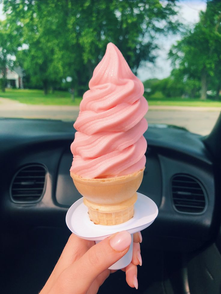 a hand holding an ice cream cone on top of a white plate in front of a car