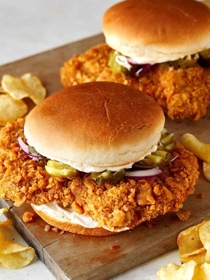 two fried chicken sandwiches sitting on top of a wooden cutting board next to potato chips