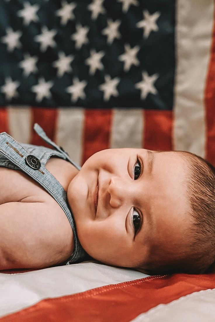 a baby laying on the ground in front of an american flag and looking at the camera