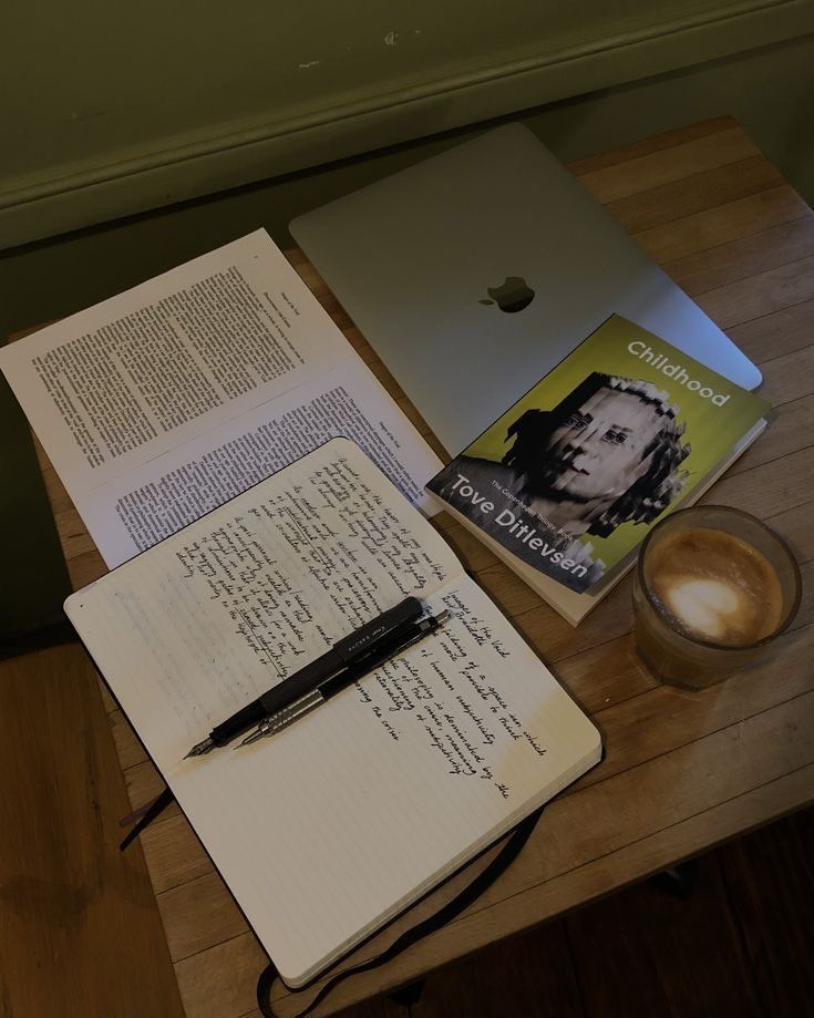 an open notebook and pen on a wooden table next to a cup of coffee with a book