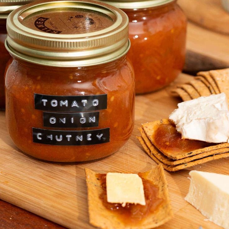 some crackers and cheese are on a cutting board next to jars of tomato chutney