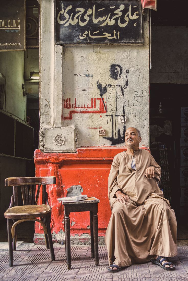 an old man sitting on a chair in front of a building with graffiti written on it