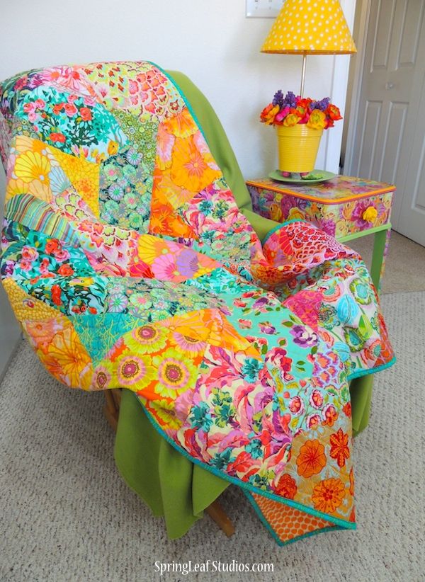 a chair with a colorful quilt on it in front of a table and lamp next to a door