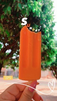 someone is holding an orange popsicle with the word sunday written on it in front of some trees