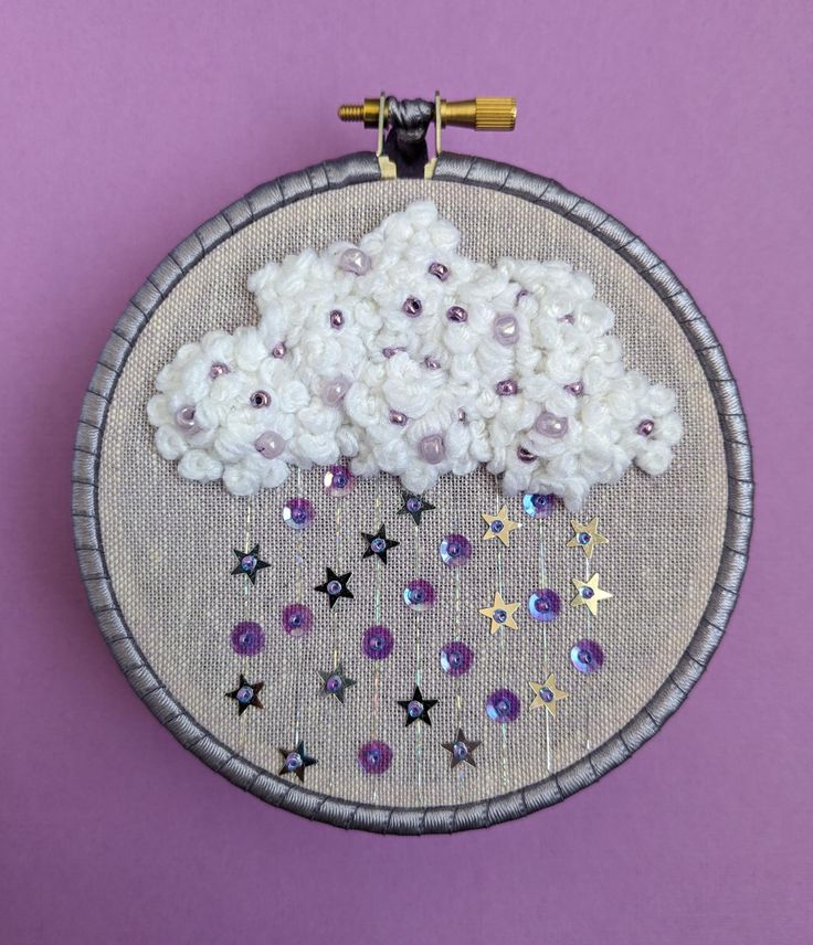 an embroidery project with white flowers and purple stars on a gray hoop hanging on a purple wall