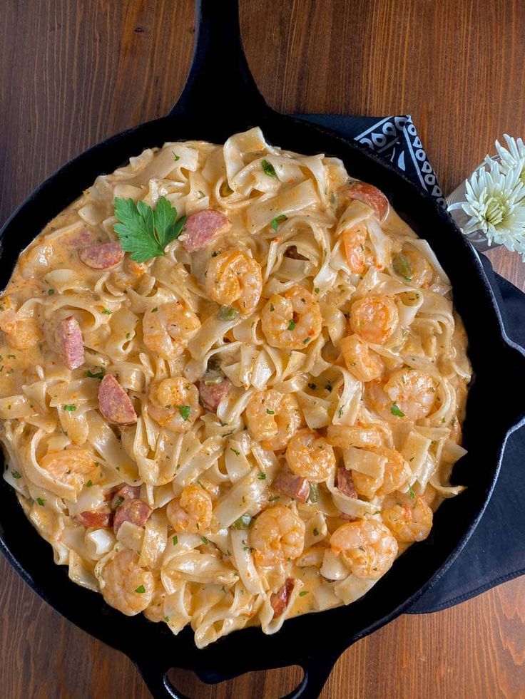 a skillet filled with pasta and shrimp on top of a wooden table next to a flower