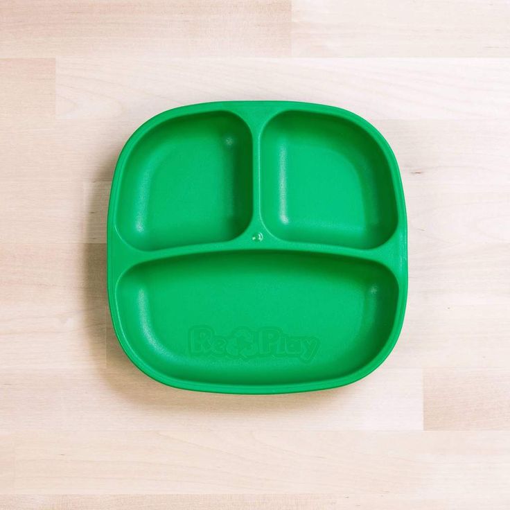 a green plastic plate with two compartments for food on top of a wooden table next to a white wall