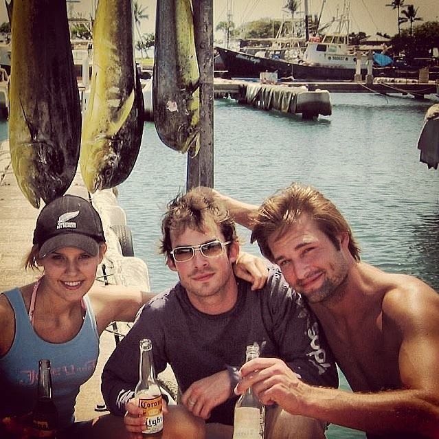 three people sitting at a table with beer bottles in front of them, posing for the camera