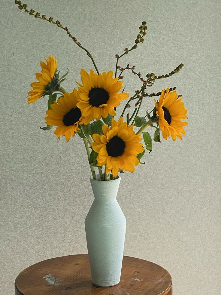 a white vase with yellow sunflowers sitting on a small wooden table next to a wall