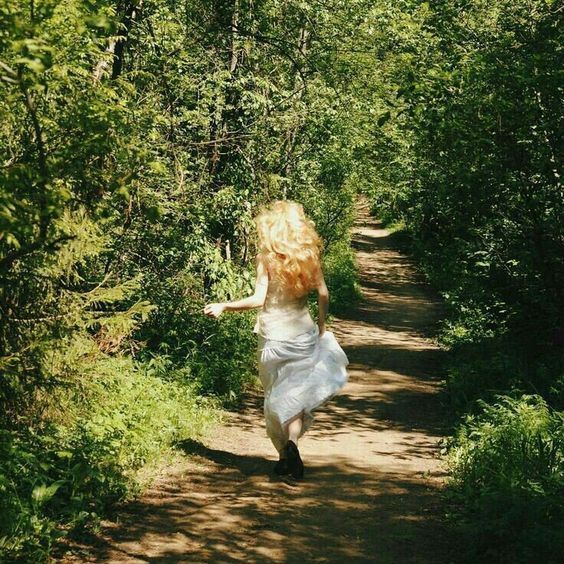 a woman in white dress walking down a dirt road surrounded by green trees and bushes