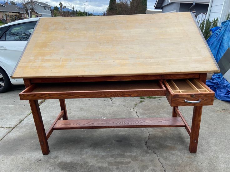 a wooden desk sitting on top of a sidewalk next to a white car and blue tarp