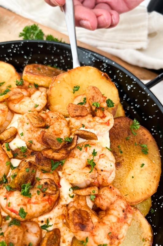 a pan filled with shrimp and potatoes on top of a wooden table next to a person's hand