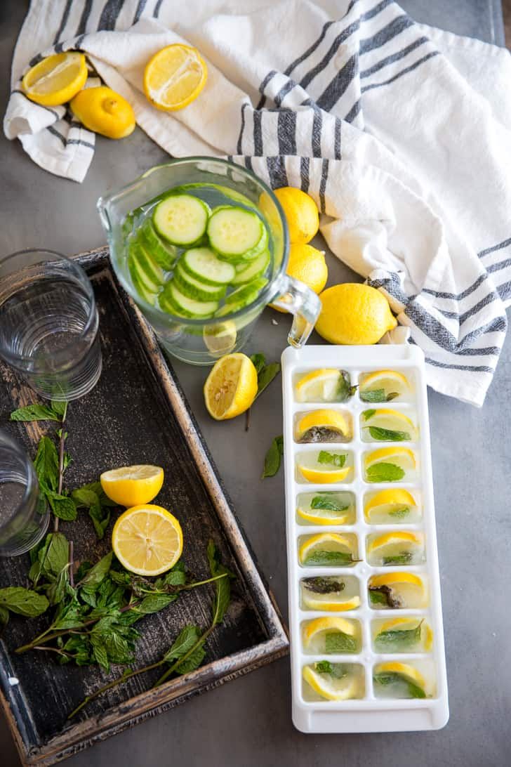 sliced lemons, cucumbers and mint leaves on a tray next to water