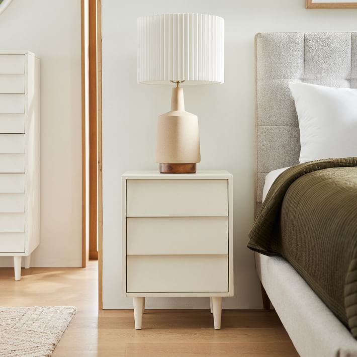 a white nightstand with a lamp on top of it next to a bed and dresser