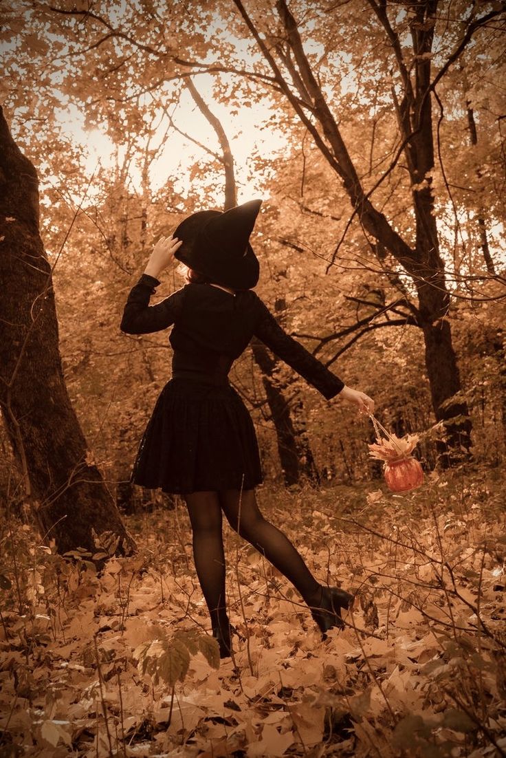 a woman in a black dress and hat walking through the woods