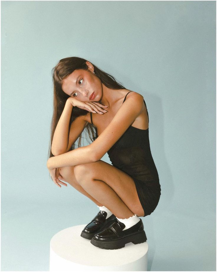 a young woman sitting on top of a white stool with her hands under her chin