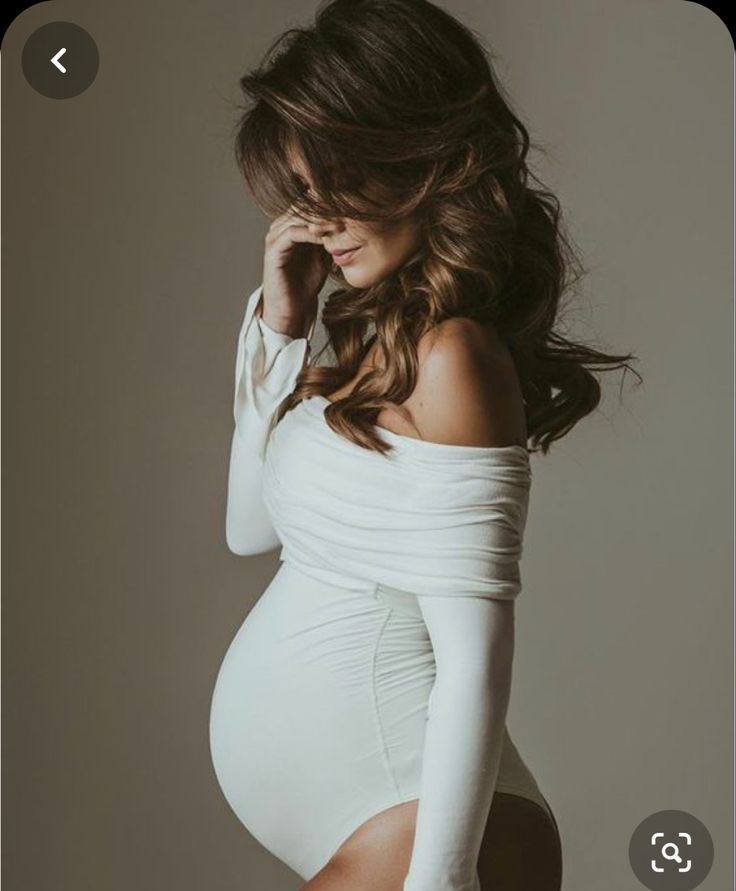 a pregnant woman in white dress posing for the camera with her hands on her head