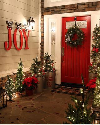 a red door with christmas decorations and lights on the front porch, surrounded by potted trees